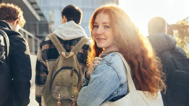 Eine junge Frau mit roten lockigen Haaren schaut in die Kamera. Im Hintergrund laufen drei junge Männer in Richtung eines Gebäudes.