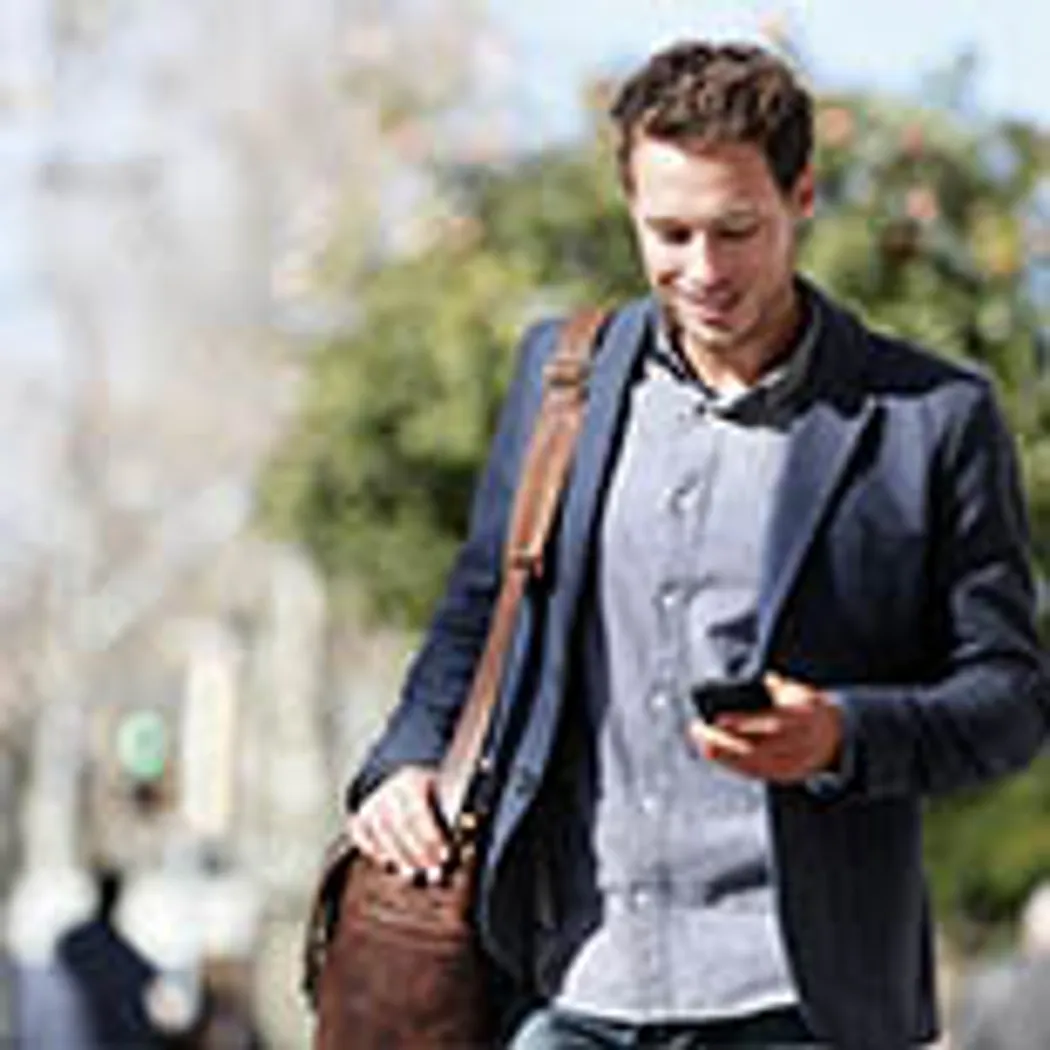 Ein Angestellter geht mit einer braunen Tasche und einem Smartphone in der Hand eine belebte Straße entlang, während er über Berufsunfähigkeitsversicherung nachdenkt. Im Hintergrund sind unscharfe Bäume sichtbar.