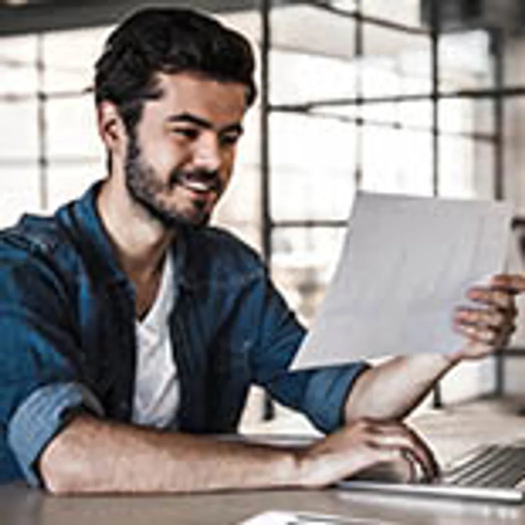 Ein junger Mann sitzt in einem modernen Büro, konzentriert auf ein Dokument. Er informiert sich über Berufsunfähigkeitsversicherung, ein wichtiges Thema für Berufsanfänger, während er am Laptop arbeitet.