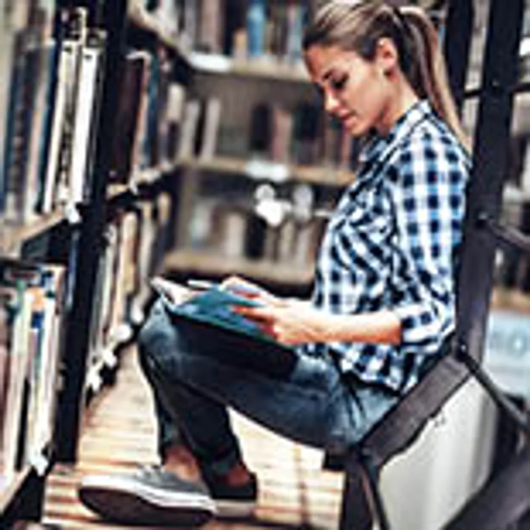 In einer Bibliothek sitzt eine junge Frau in Freizeitkleidung auf dem Boden und liest ein Buch. Sie könnte eine Schülerin oder Studentin sein, die sich über Berufsunfähigkeitsversicherung informiert. Im Hintergrund sind Bücherregale sichtbar.