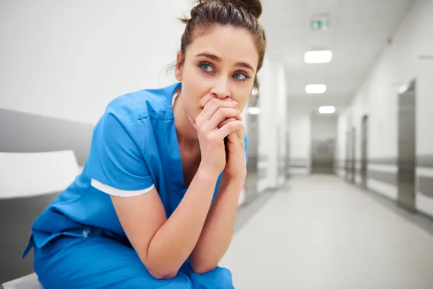 Eine besorgte Krankenschwester in blauer Arbeitskleidung sitzt auf einem Stuhl im hellen Krankenhausflur, die Hände vor dem Gesicht verschränkt. Der Flur erstreckt sich in die Ferne.