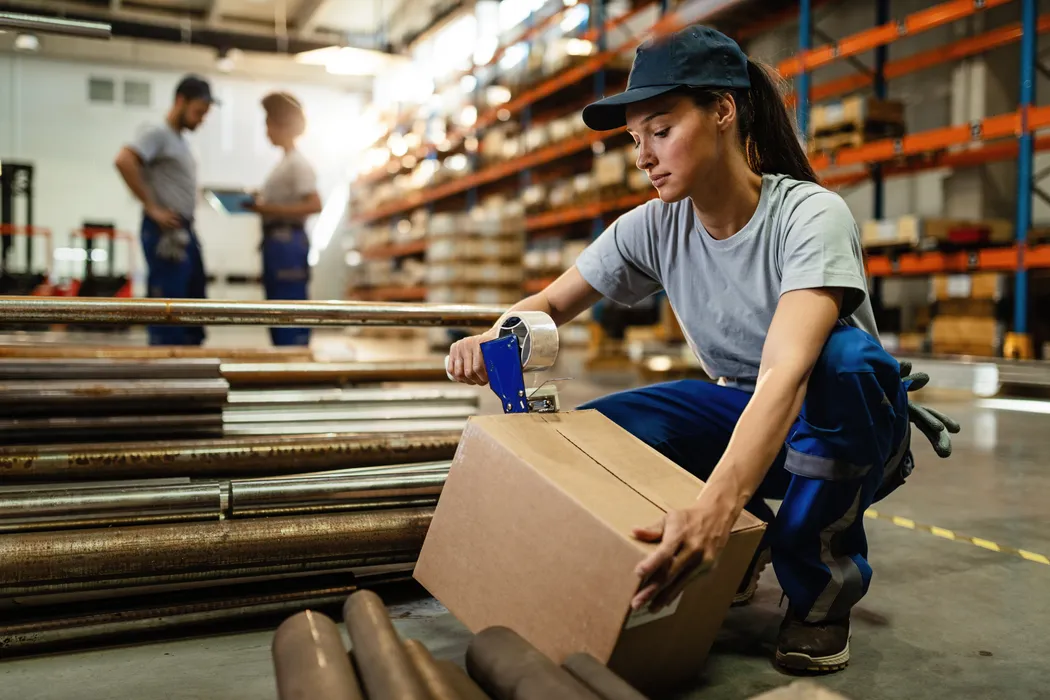 Frau in Arbeitskleidung kniet im Lager und verklebt einen Karton für den Versand. In einem Lager kniet eine Frau in Arbeitskleidung und verschließt einen Karton mit Klebeband. Im Hintergrund sind Regale und zwei Personen in Gespräch vertieft. Der Karton wird für den Versand vorbereitet.