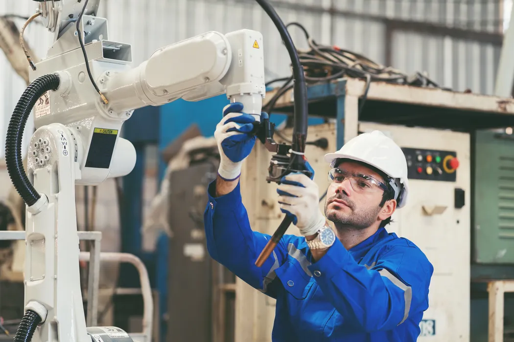 Arbeiter in blauer Uniform und weißem Schutzhelm justiert in einer Industrieanlage einen Roboterarm. Ein Arbeiter in blauer Uniform und weißem Schutzhelm bedient einen Roboterarm in einer Industrieanlage, wobei er präzise Einstellungen vornimmt.