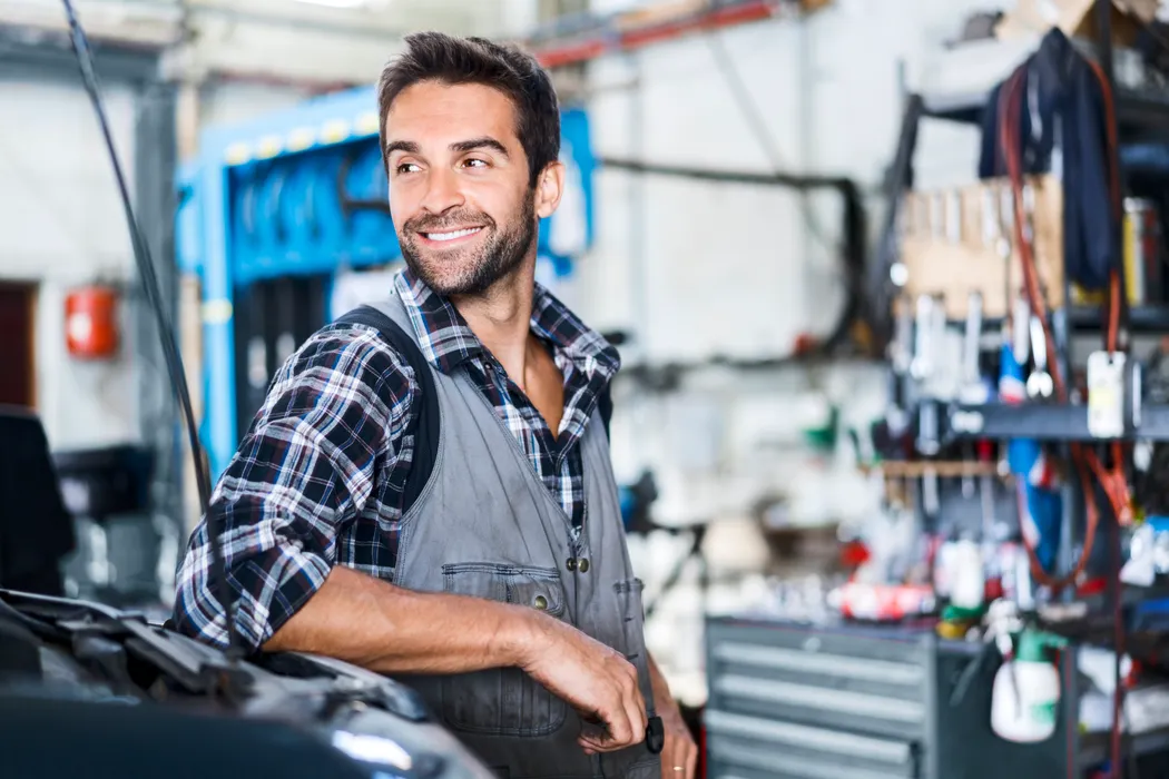 Handwerker in Arbeitskleidung lehnt in einer Werkstatt an der Motorhaube eines Autos. In einer gut ausgestatteten Werkstatt lehnt ein Handwerker in Arbeitskleidung entspannt an der geöffneten Motorhaube eines Autos, umgeben von Werkzeugen und Ersatzteilen.