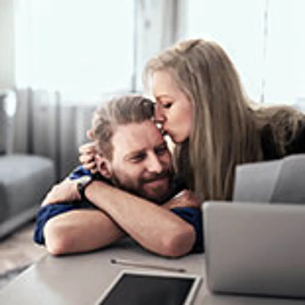Risikolebensversicherung: Lebenspartner In einem hellen Raum sitzt ein Paar an einem Tisch, umgeben von Laptop und Tablet. Der Lebenspartner legt beruhigend den Arm um die andere Person, während sie über Risikolebensversicherung sprechen.