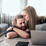 In einem hellen Raum sitzt ein Paar an einem Tisch, umgeben von Laptop und Tablet. Der Lebenspartner legt beruhigend den Arm um die andere Person, während sie über Risikolebensversicherung sprechen.