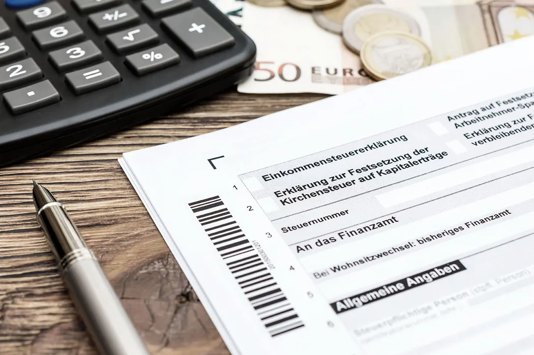 Sterbegeldversicherung und Steuer Eine Steuererklärung auf einem Holztisch, flankiert von einem Kugelschreiber und einem Taschenrechner. Im Hintergrund liegen Münzen und ein 50-Euro-Schein. Eine Sterbegeldversicherung wird nicht direkt dargestellt, könnte aber im Kontext der finanziellen Planung relevant sein.