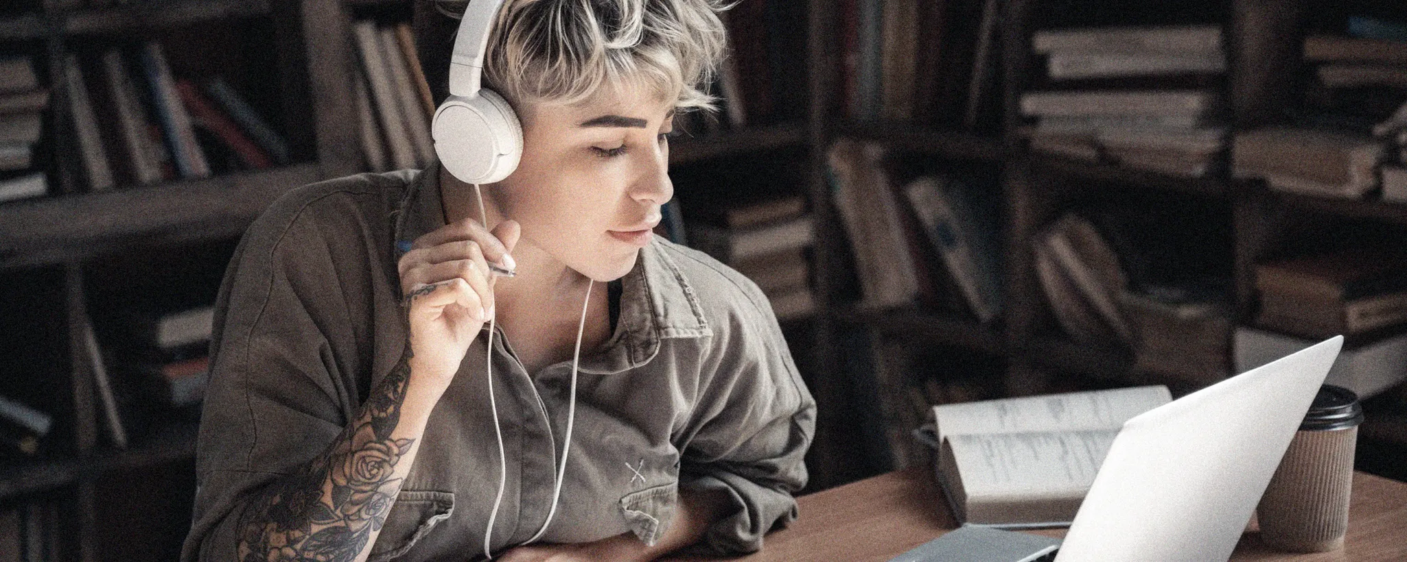 Eine Person sitzt an einem Holztisch in einem gemütlichen Raum mit Bücherregalen im Hintergrund, die mit verschiedenen Büchern gefüllt sind. Sie trägt große, weiße Kopfhörer und hat eine tätowierte Hand, die nachdenklich an ihrem Kinn ruht. Die Person trägt ein olivgrünes Hemd, das gut zu ihrer entspannten Haltung passt. Auf dem Tisch vor ihr steht ein aufgeklappter Laptop, der für Arbeit oder Studium genutzt wird, und daneben ein aufgeschlagenes Notizbuch, das mit handschriftlichen Notizen gefüllt ist. Rechts vom Laptop befindet sich ein Pappbecher, vermutlich für Kaffee, der auf eine Pause oder kreative Anregung hinweist.
