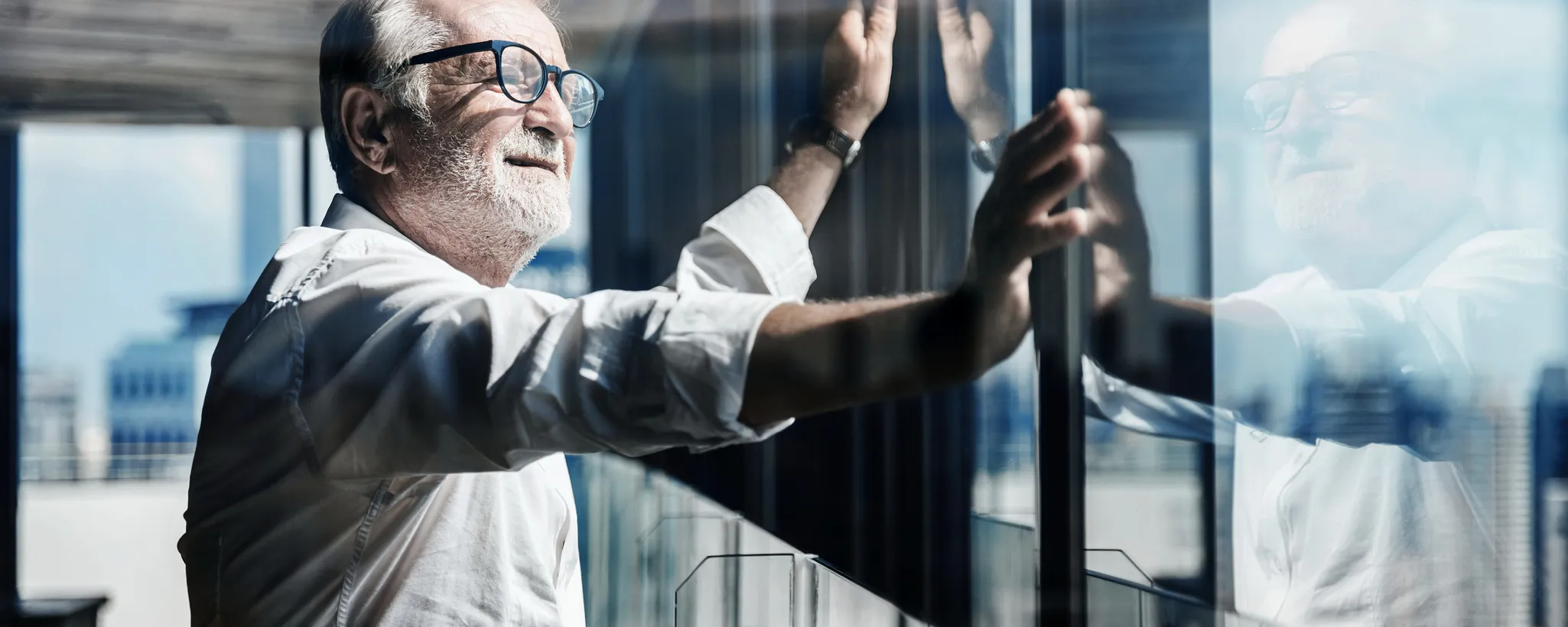 Ein Mann in einem weißen Hemd steht an einem Fenster und blickt nach draußen. Die Architektur des Gebäudes spiegelt sich in der Scheibe. Themen wie Meldepflicht und Krankenversicherung könnten in diesem Kontext relevant sein.