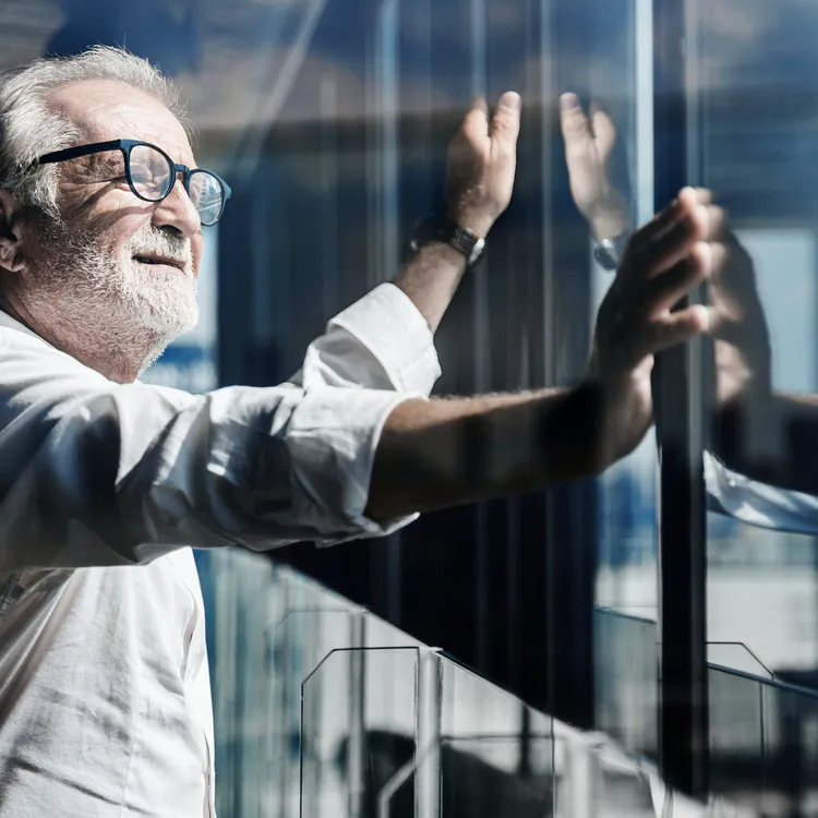 Ein Mann in einem weißen Hemd steht an einem Fenster und blickt nach draußen. Die Architektur des Gebäudes spiegelt sich in der Scheibe. Themen wie Meldepflicht und Krankenversicherung könnten in diesem Kontext relevant sein.