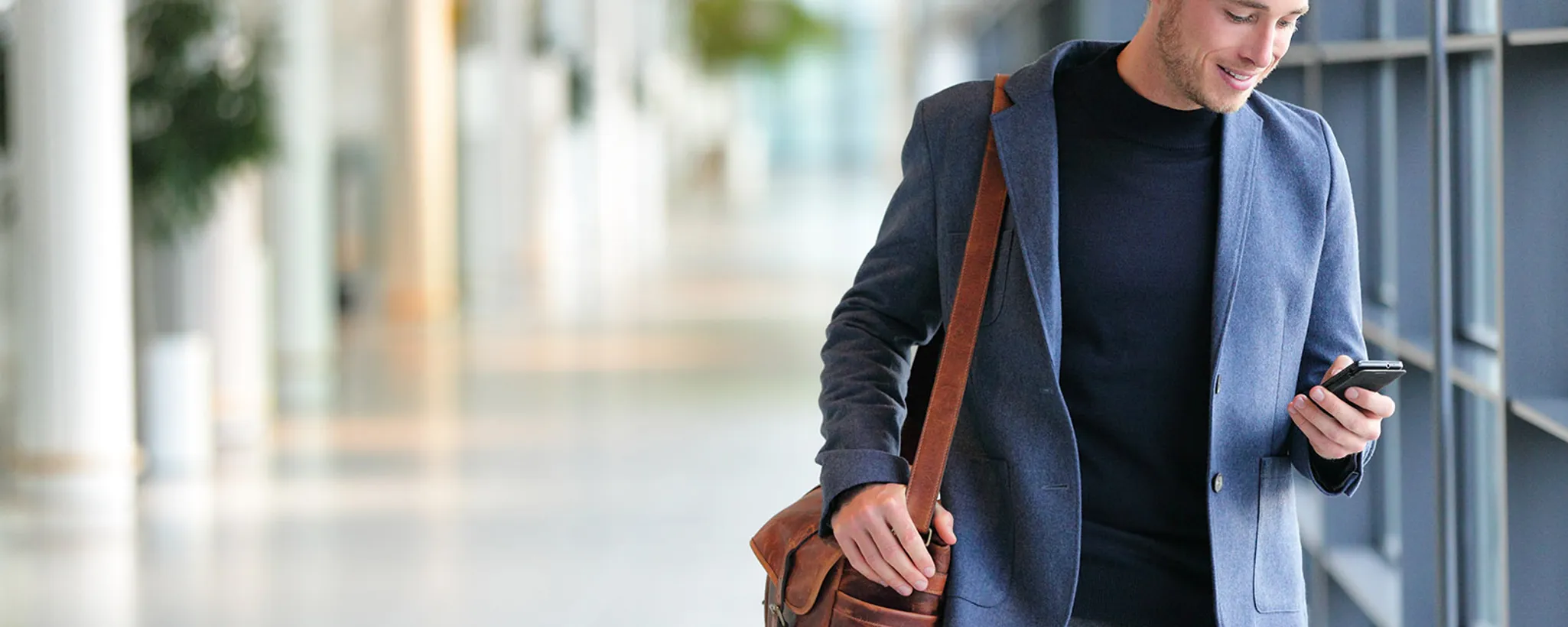 Ein Mann in einem eleganten blauen Blazer geht durch einen modernen Büroflur, eine Ledertasche über der Schulter und ein Smartphone in der Hand. Er könnte sich mit Berufsunfähigkeitsversicherung oder Leistungsfällen beschäftigen.