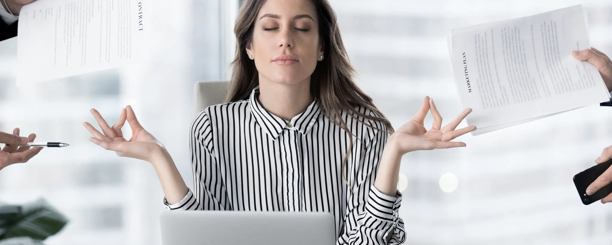 Eine Frau sitzt an einem Tisch, vor ihr ein geöffneter Laptop, der eine wichtige Rolle in ihrem Arbeitsalltag spielt. Sie trägt ein schwarz-weiß gestreiftes Hemd, das einen professionellen Eindruck vermittelt. Ihre Hände sind in einer meditativen Haltung mit Daumen und Zeigefinger zusammengeführt, was auf ihre Fähigkeit zur Konzentration hinweist. Um sie herum sind mehrere Hände zu sehen, die ihr Dokumente und einen Stift entgegenstrecken, als ob sie ihre Aufmerksamkeit erfordern, was den Druck und die Anforderungen ihrer Umgebung verdeutlicht. Der Hintergrund ist verschwommen und zeigt ein helles, modernes Bürogebäude, das die dynamische und geschäftige Atmosphäre unterstreicht. Die Szene vermittelt
