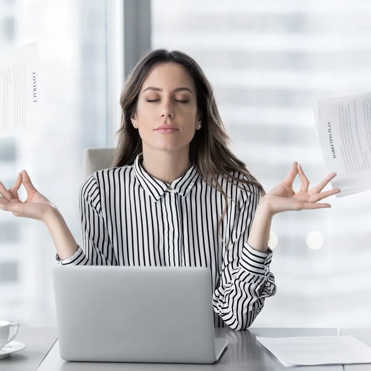 Eine Frau sitzt an einem Tisch, vor ihr ein geöffneter Laptop, der eine wichtige Rolle in ihrem Arbeitsalltag spielt. Sie trägt ein schwarz-weiß gestreiftes Hemd, das einen professionellen Eindruck vermittelt. Ihre Hände sind in einer meditativen Haltung mit Daumen und Zeigefinger zusammengeführt, was auf ihre Fähigkeit zur Konzentration hinweist. Um sie herum sind mehrere Hände zu sehen, die ihr Dokumente und einen Stift entgegenstrecken, als ob sie ihre Aufmerksamkeit erfordern, was den Druck und die Anforderungen ihrer Umgebung verdeutlicht. Der Hintergrund ist verschwommen und zeigt ein helles, modernes Bürogebäude, das die dynamische und geschäftige Atmosphäre unterstreicht. Die Szene vermittelt