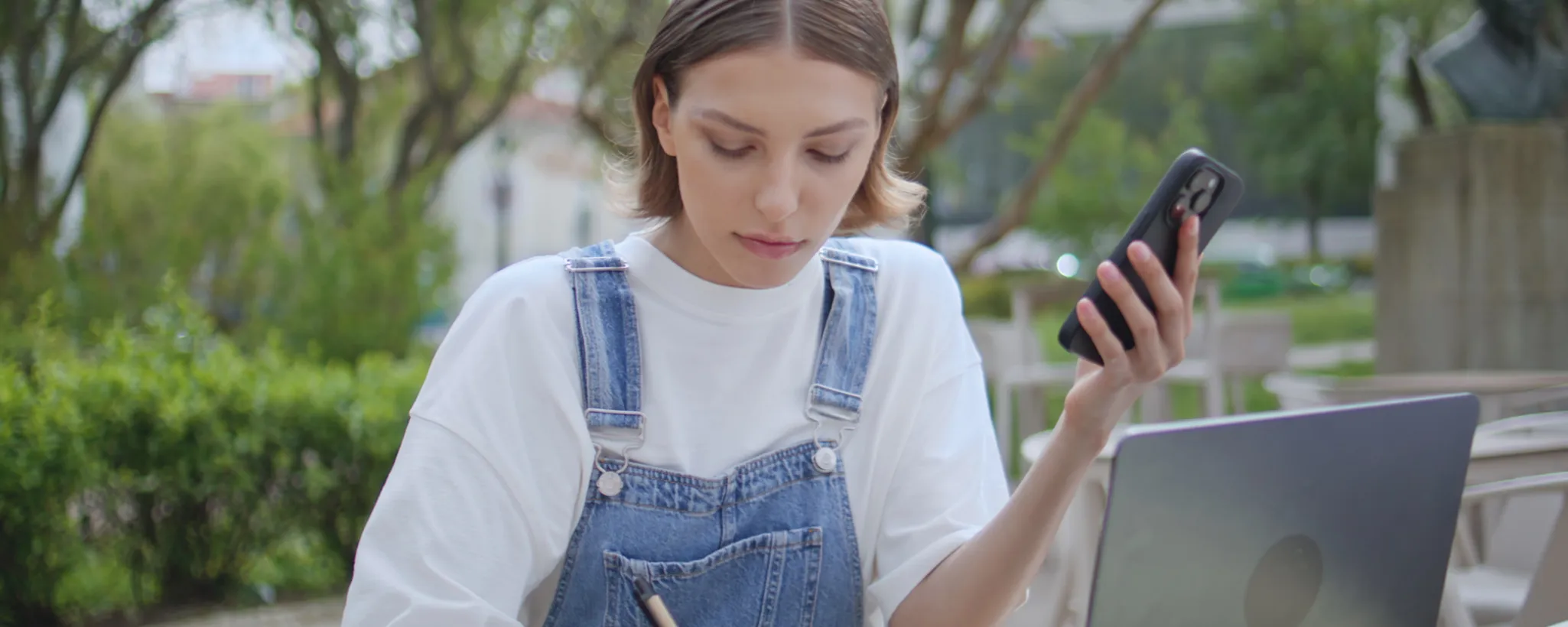 Eine junge Frau sitzt in einem Park vor ihrem Laptop. Sie hält ein Smartphone in der Hand und schreibt mit einem Stift Informationen auf, anhand welcher sie sich einen Lebensplan erstellen kann.