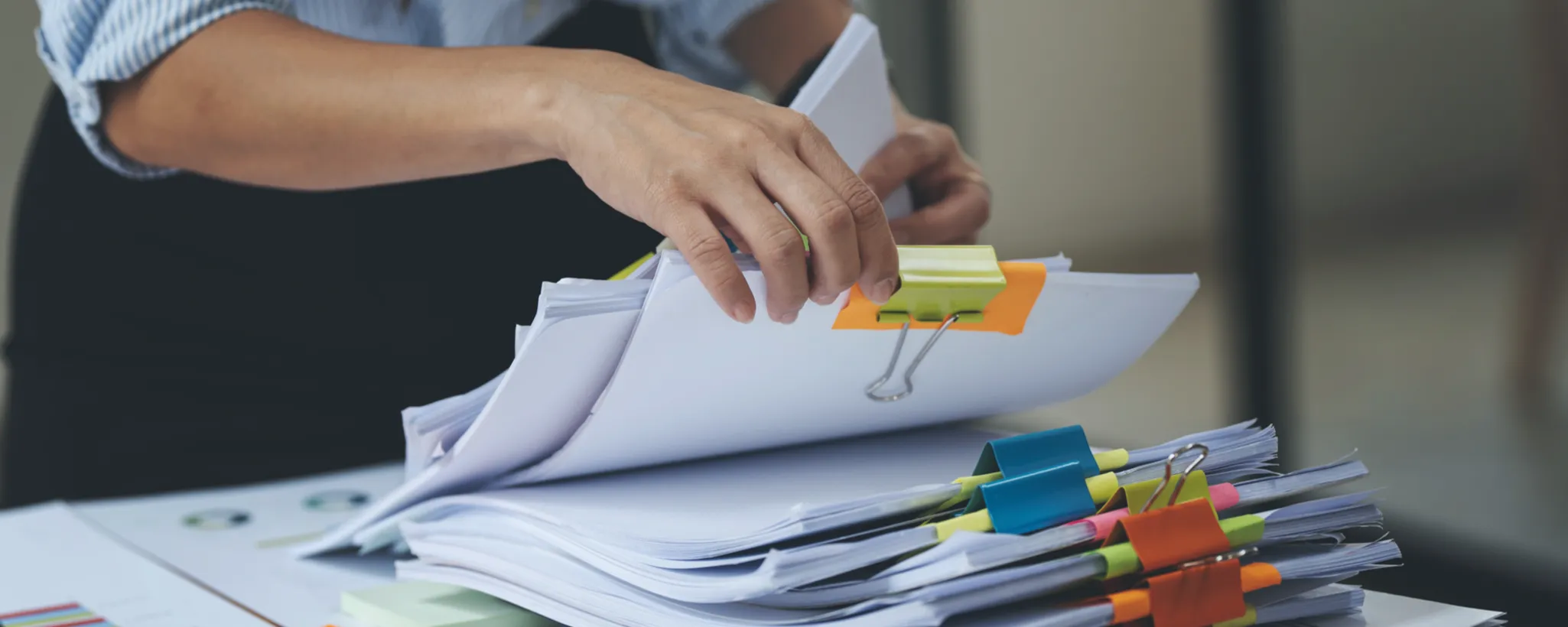 Eine Hand sortiert Papierstapel im Büro, die mit bunten Klammern organisiert sind. Diese ordentliche Struktur fördert die Produktivität durch klare und übersichtliche Dokumentenverwaltung.
