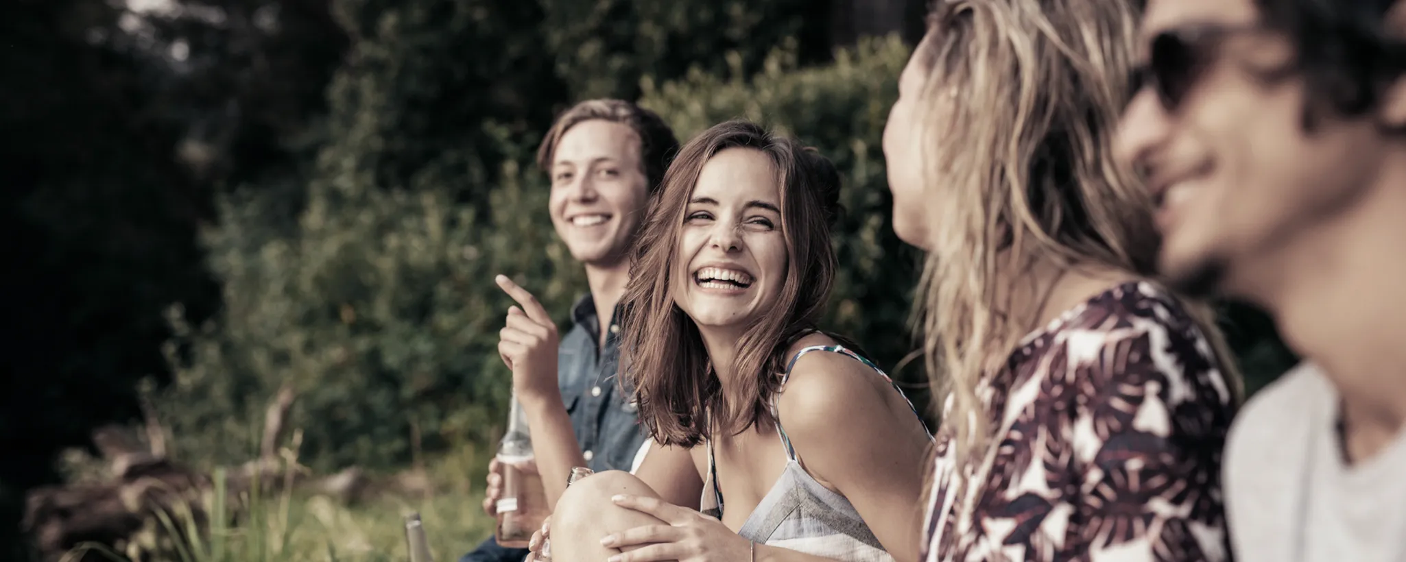 Eine Gruppe von Menschen sitzt im Freien und führt lebhafte Gespräche. Im Vordergrund sind grüne Pflanzen zu sehen, während die Personen sich über Themen wie Studium und Unterhalt unterhalten.