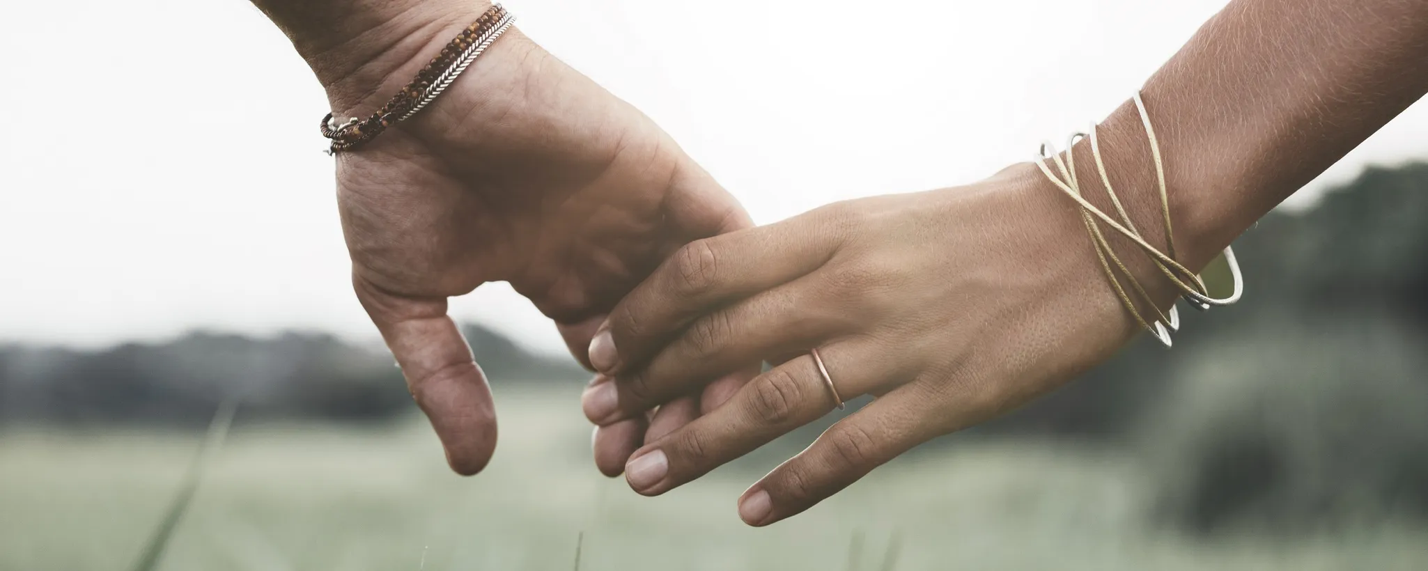 Zwei Hände, geschmückt mit Armbändern, greifen sich sanft in einer grünen Landschaft. Diese Geste symbolisiert Vertrauen und Nähe, ähnlich einer wilden Ehe, die ohne formellen Bund besteht. Die Freiheit und Ungezwungenheit einer solchen Beziehung wird durch die natürliche Umgebung und die entspannte Haltung der Hände unterstrichen, die die Essenz einer wilden Ehe verkörpern.