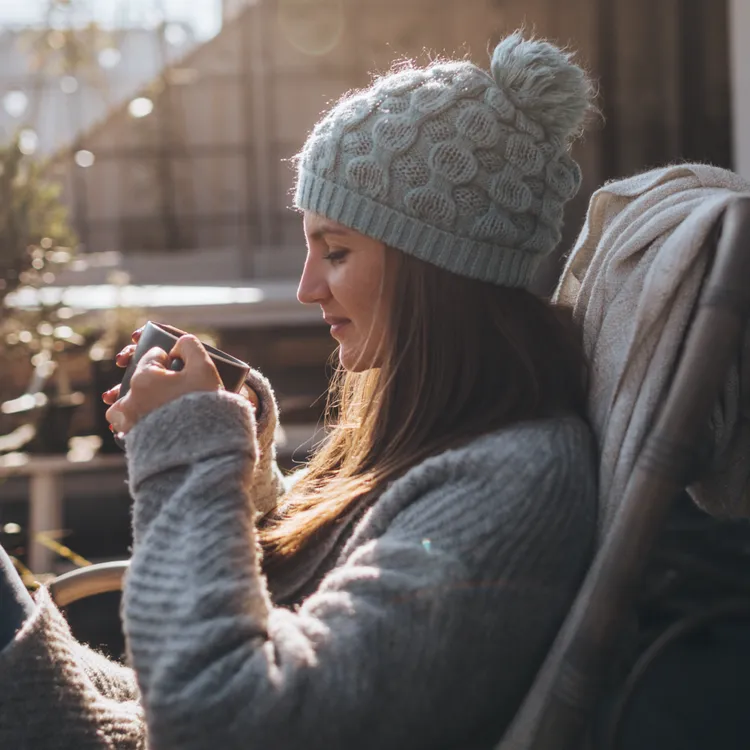 Eine alleinerziehende Person sitzt entspannt in einem gemütlichen Sessel auf der Terrasse ihres Hauses, genießt die Sonne und hält ein Smartphone in der Hand. Sie trägt warme Kleidung und wirkt zufrieden in ihrem Zuhause.