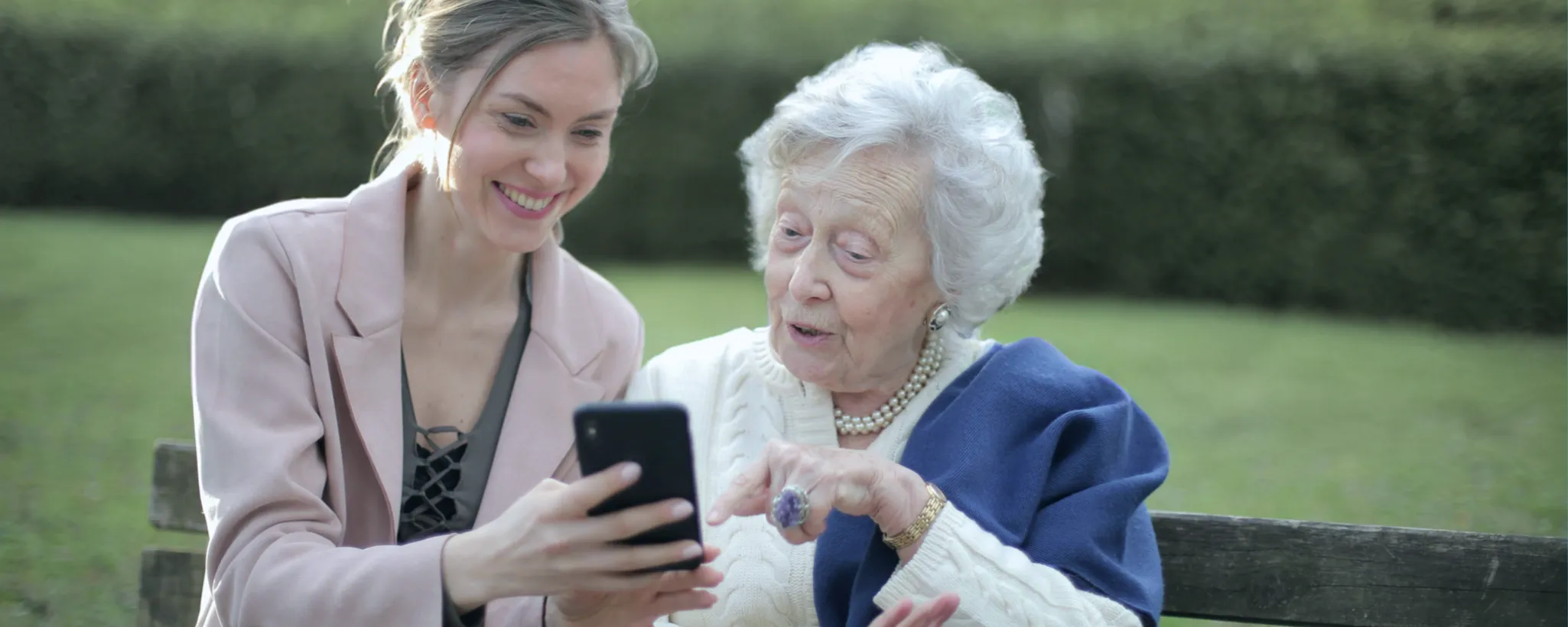 Zwei Frauen sitzen auf einer Parkbank, während die ältere Frau interessiert auf ein Smartphone schaut, das die jüngere Frau hält. Sie diskutieren über einen digitalen Grabstein. Im Hintergrund ist eine grüne Hecke zu sehen.