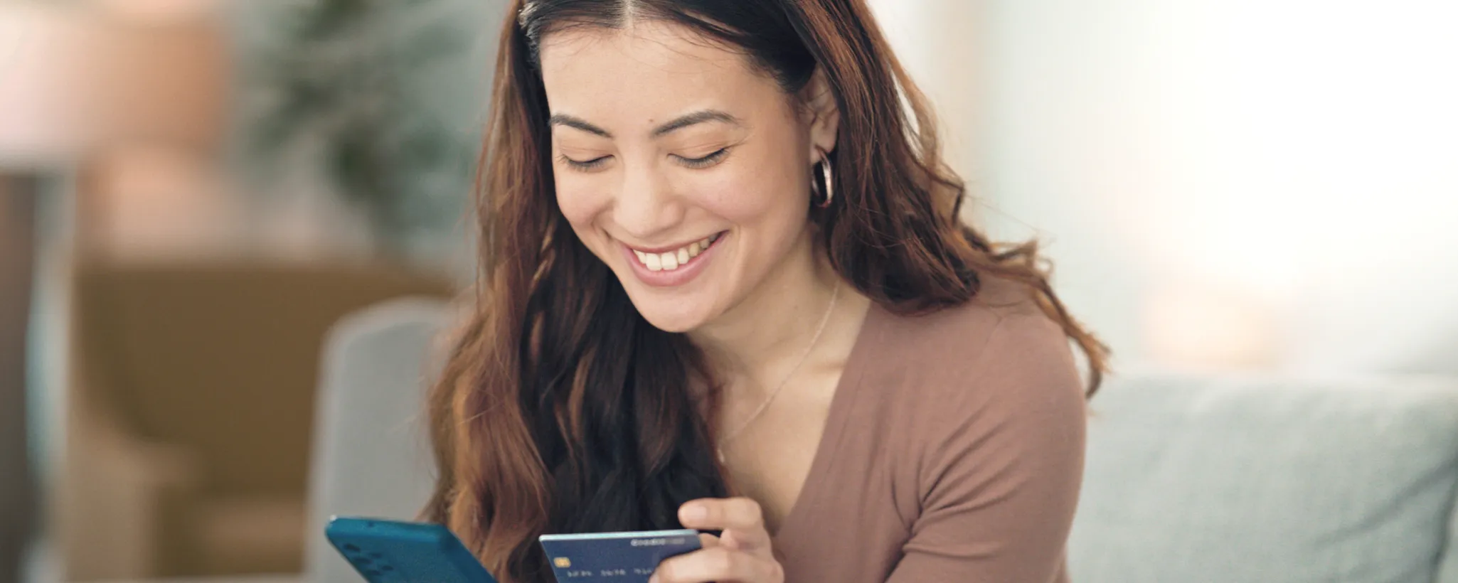 Eine Frau sitzt entspannt auf einem Sofa und hält ein Smartphone sowie eine Kreditkarte in den Händen, symbolisierend finanzielle Unabhängigkeit im digitalen Zeitalter.