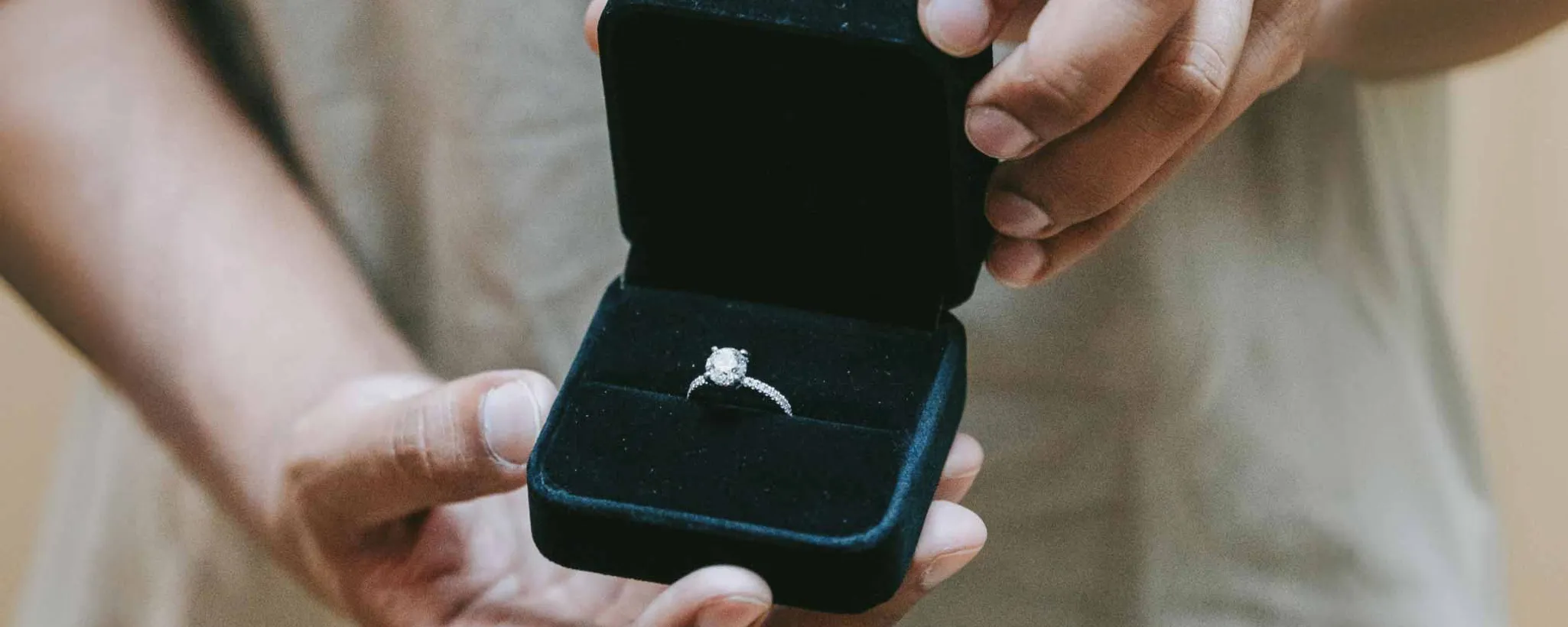 Eine Person zeigt einem anderen Menschen ein geöffnetes Ringetui mit einem Verlobungsring. Dabei sind nur die Hände der einen Person zu sehen.