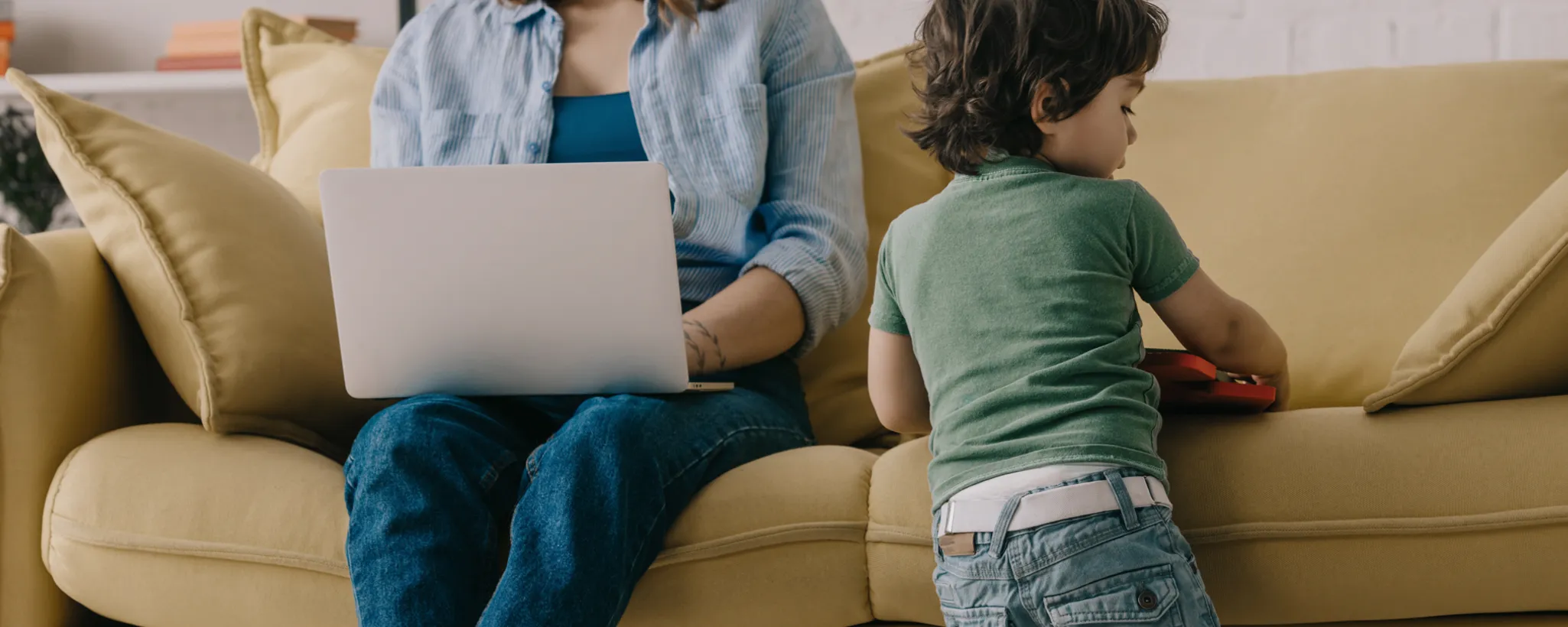 Eine Person arbeitet mit einem Laptop auf einem Sofa, während ein Kind daneben steht und spielt. Die Szene symbolisiert den Wiedereinstieg nach der Elternzeit und gibt Tipps für einen erfolgreichen Jobwechsel.
