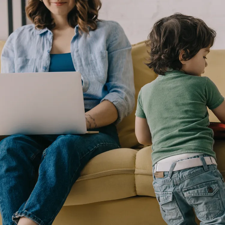 Eine Person arbeitet mit einem Laptop auf einem Sofa, während ein Kind daneben steht und spielt. Die Szene symbolisiert den Wiedereinstieg nach der Elternzeit und gibt Tipps für einen erfolgreichen Jobwechsel.
