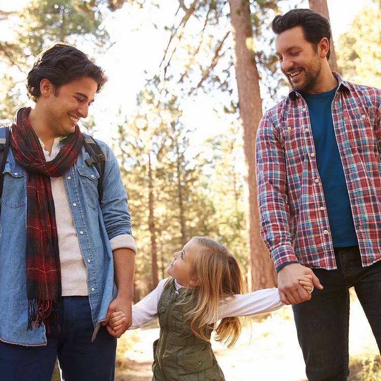 Zwei Männer, leger gekleidet in Jeans und Hemden, stehen in einem Wald und halten die Hände eines fröhlichen Kindes. Der Hauptverdiener der Familie scheint das Kind liebevoll zu führen.