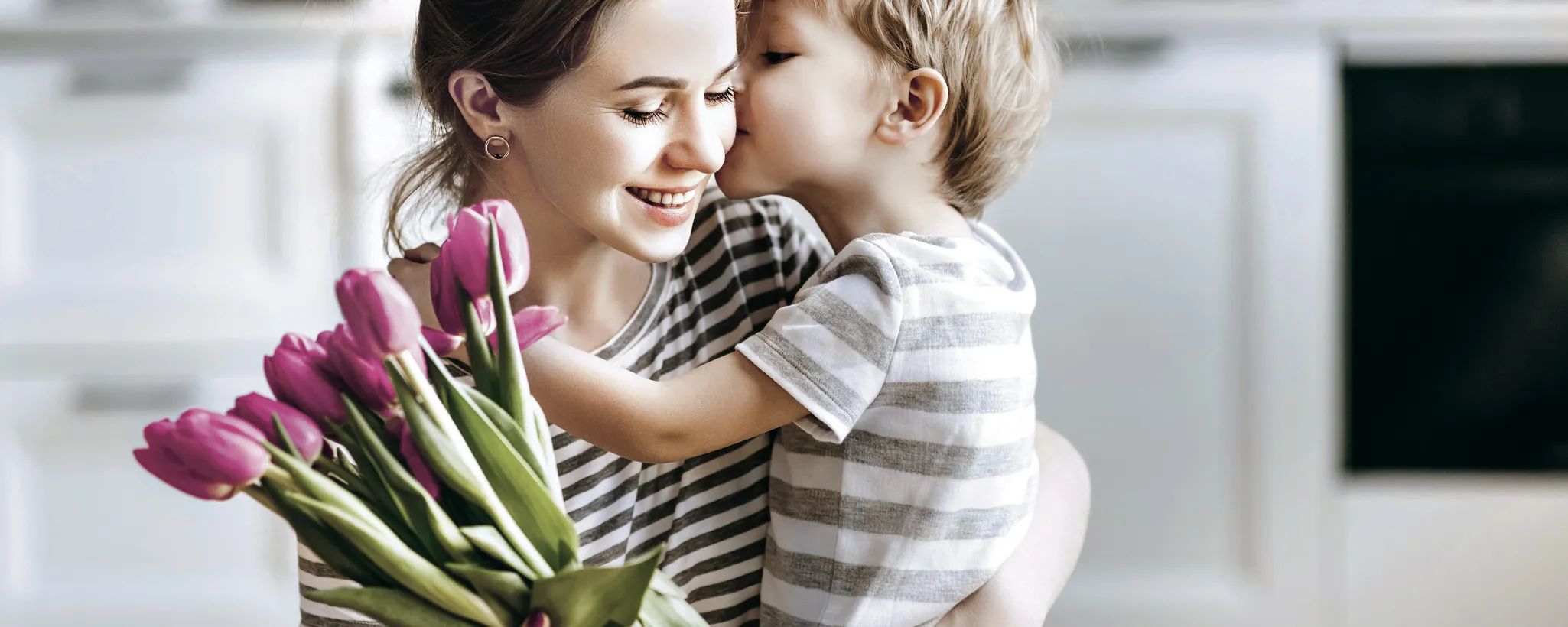 In einer modernen Küche hält eine Frau einen kleinen Jungen im Arm, während sie einen Strauß rosa Tulpen überreicht bekommt, was die Gegenwart des Muttertags symbolisiert und dessen Entstehung feiert.