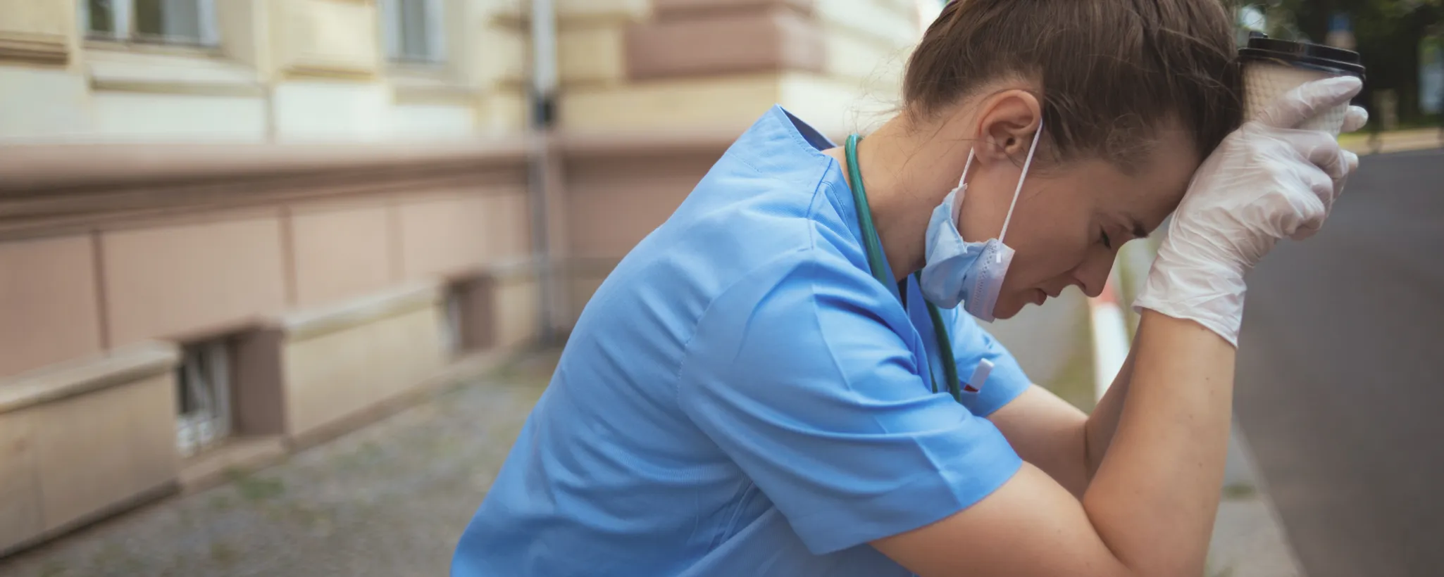 Eine Pflegekraft in blauer Uniform sitzt erschöpft auf einer Bank, den Kopf auf ihre Arme gestützt. Sie trägt Handschuhe und hält einen Kaffeebecher. Die Szene vermittelt Gedanken über die Zukunft der Pflegeberufe.