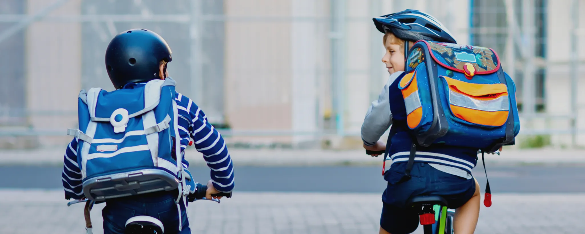 Zwei Grundschulkinder mit Schulranzen fahren mit dem Fahrrad. Sie sind von hinten zu sehen und wirken fröhlich und voller Vorfreude auf die Schule