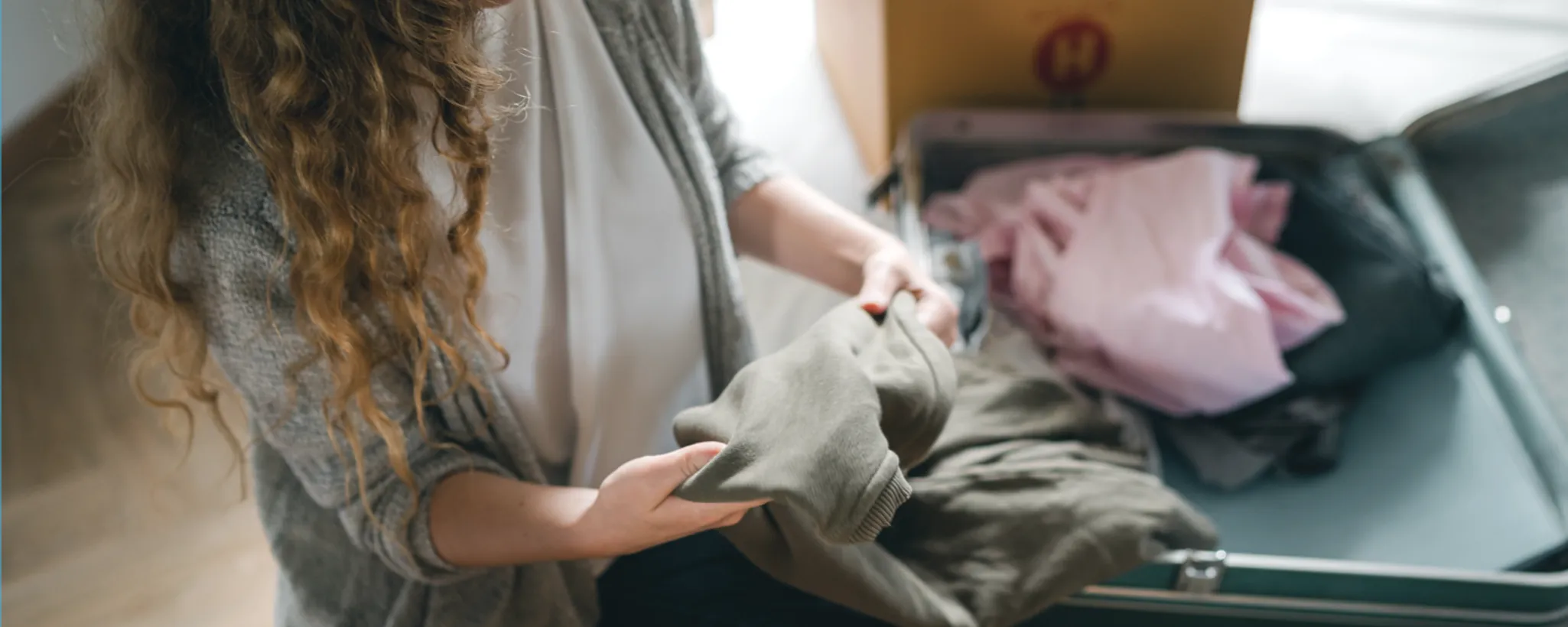 Eine Frau sitzt vor einem geöffneten Koffer und hält ein grünes Kleidungsstück in den Händen. Neben ihr liegen weitere Kleidungsstücke, darunter ein rosa Kindershirt. Im Hintergrund steht ein Karton.