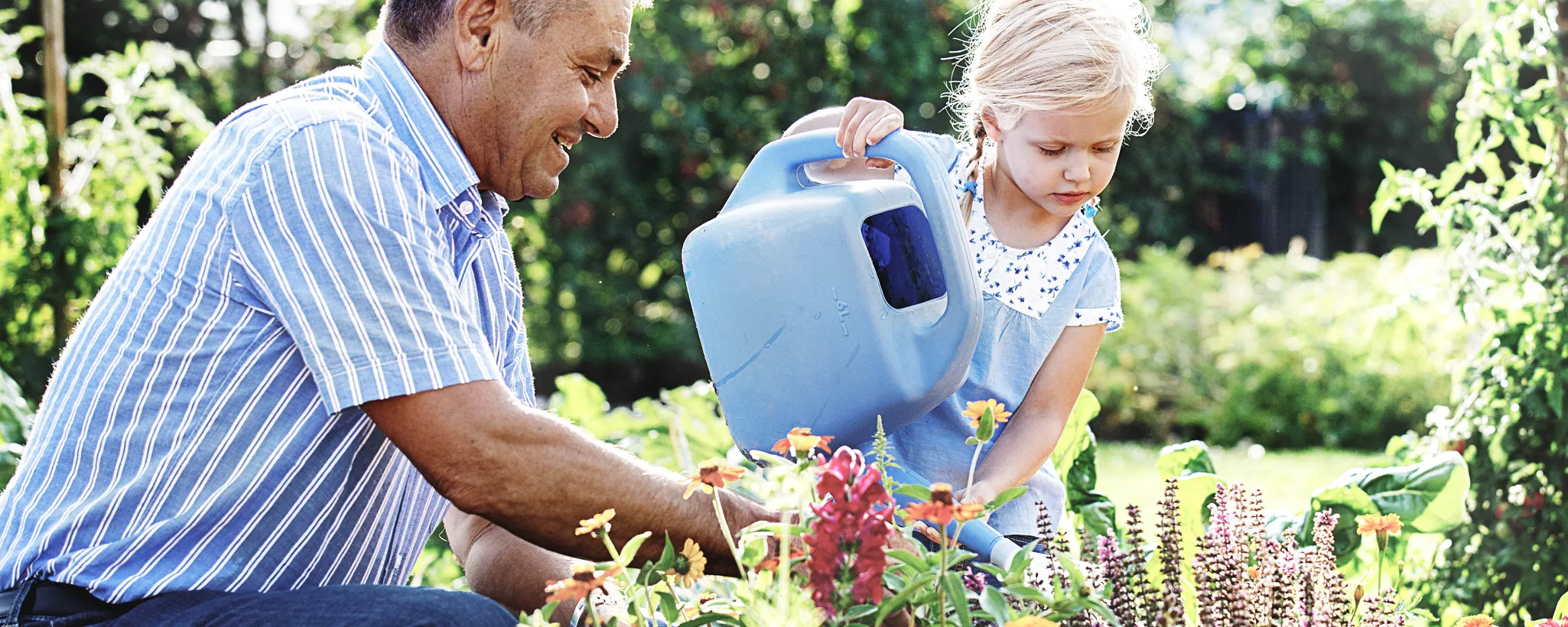 Ein Kind gießt mit einer blauen Kanne bunte Blumen in einem gepflegten Garten, während ein Erwachsener beim Frühjahrsputz die Pflanzen sorgfältig pflegt. Die Szene zeigt harmonische Gartenarbeit.
