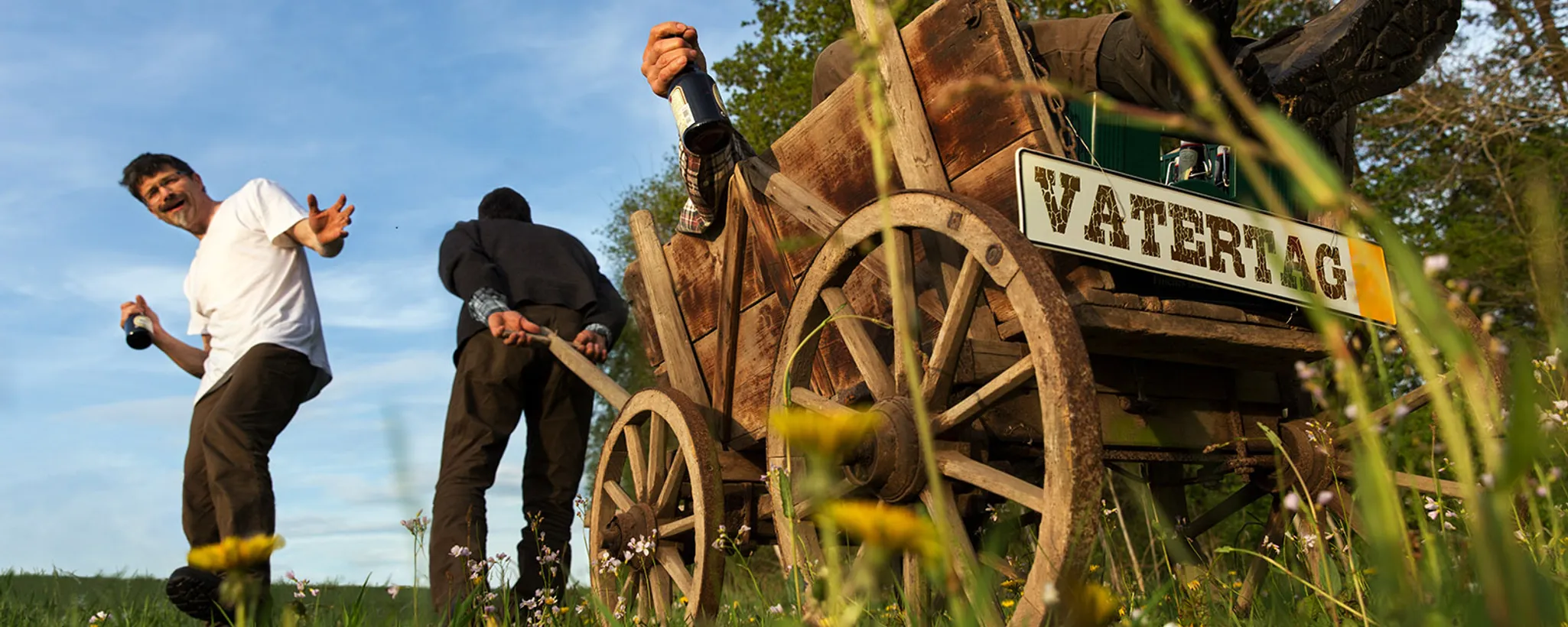 Auf einer grünen Wiese ziehen zwei Männer einen hölzernen Bollerwagen, auf dem ein Schild mit der Aufschrift "Vatertag" angebracht ist. Einer der Männer hält eine Flasche, was an einen ausgelassenen Unfalltag erinnert.