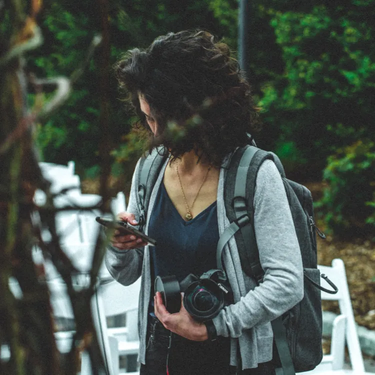 Eine Frau im Freien, möglicherweise im Urlaub, hält ihre Kamera und schaut auf ihr Handy. Sie trägt einen Rucksack, um sich vor Diebstahl zu schützen, während sie durch den Park geht. Die leeren weißen Stühle im Hintergrund könnten von einem kürzlich stattgefundenen Ereignis stammen, das keinen sichtbaren Schaden hinterlassen hat.