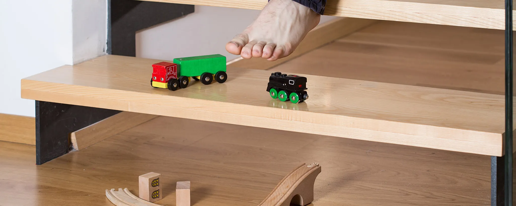 Auf einer hölzernen Treppe im Zuhause sind Spielzeugzüge platziert, während ein Fuß auf der Stufe ruht. Der Ort wirkt wie ein kreativer Spielplatz in der kindlichen Welt.