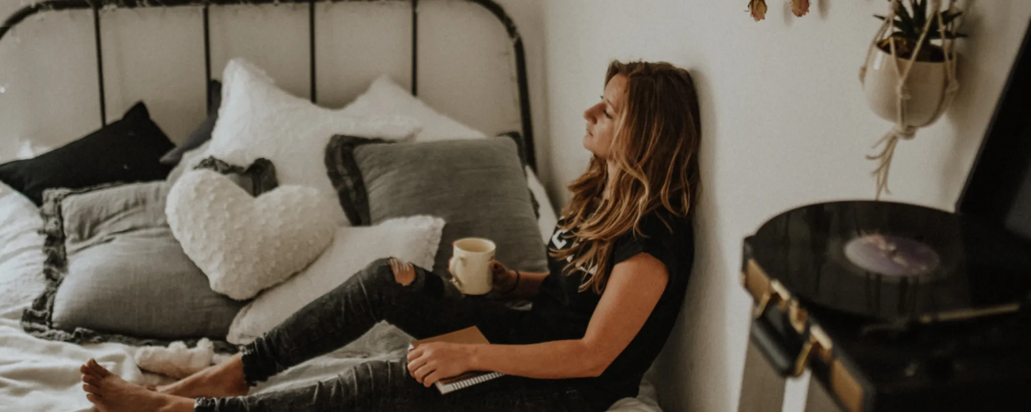 Eine Frau sitzt entspannt mit einer Tasse Kaffee auf einem Bett, umgeben von Kissen. Neben ihr liegt ein Notizbuch, möglicherweise zum Thema Krankengeld. Im Vordergrund steht ein Plattenspieler.