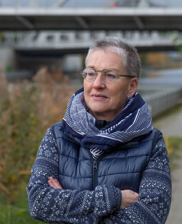 Gudrun Jay-Bößl, eine erfahrene Heilpraktikerin für Psychotherapie, steht mit verschränkten Armen in einem Wintermantel und Schal vor einer Brücke, umgeben von herbstlicher Landschaft.