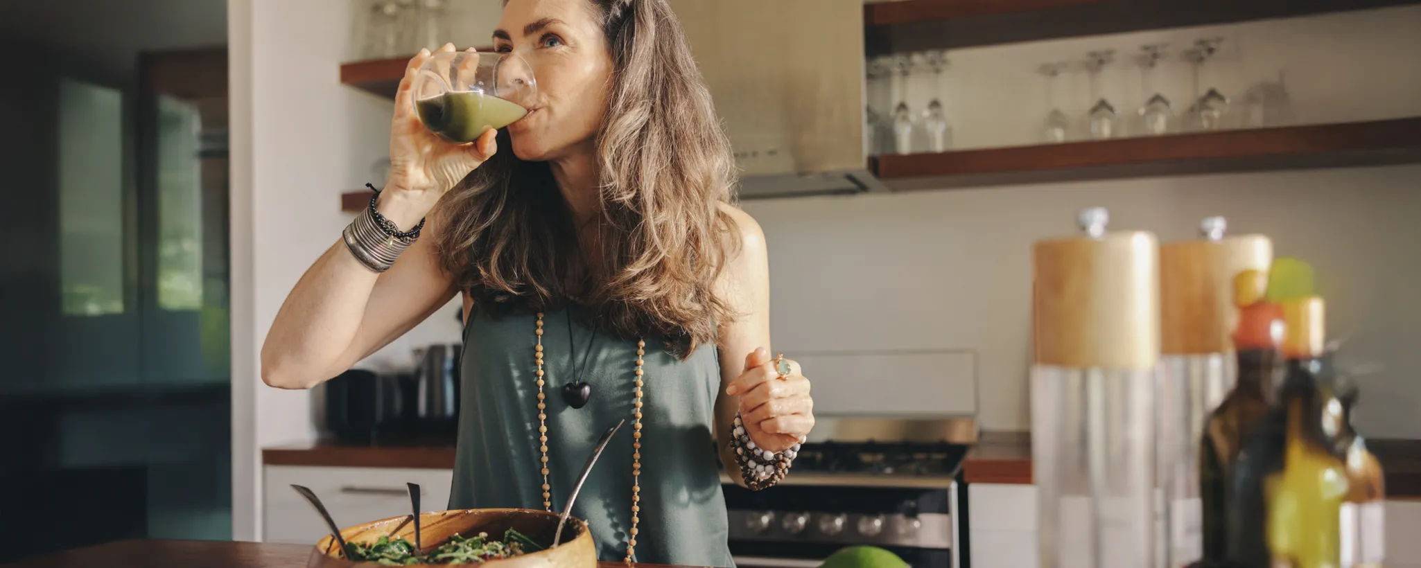 In einer modernen Küche steht eine Frau, die einen grünen Smoothie trinkt und eine Schüssel mit frischem Salat zubereitet, was auf eine bewusste Ernährung hinweist. Die ruhige Umgebung fördert eine positive Psyche.