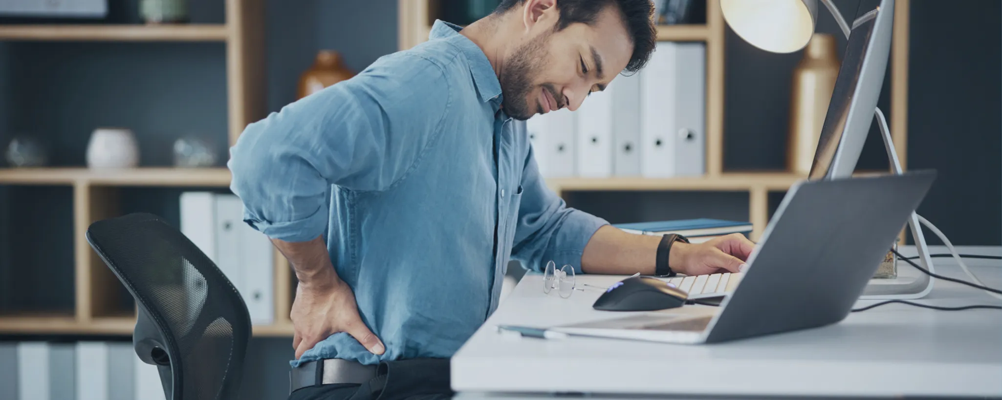 Ein Mann sitzt an einem Bürotisch vor einem Computer und hält sich den Rücken. Der Arbeitsplatz ist mit Regalen im Hintergrund ausgestattet. Ein gesunder Rücken scheint ihm wichtig zu sein.