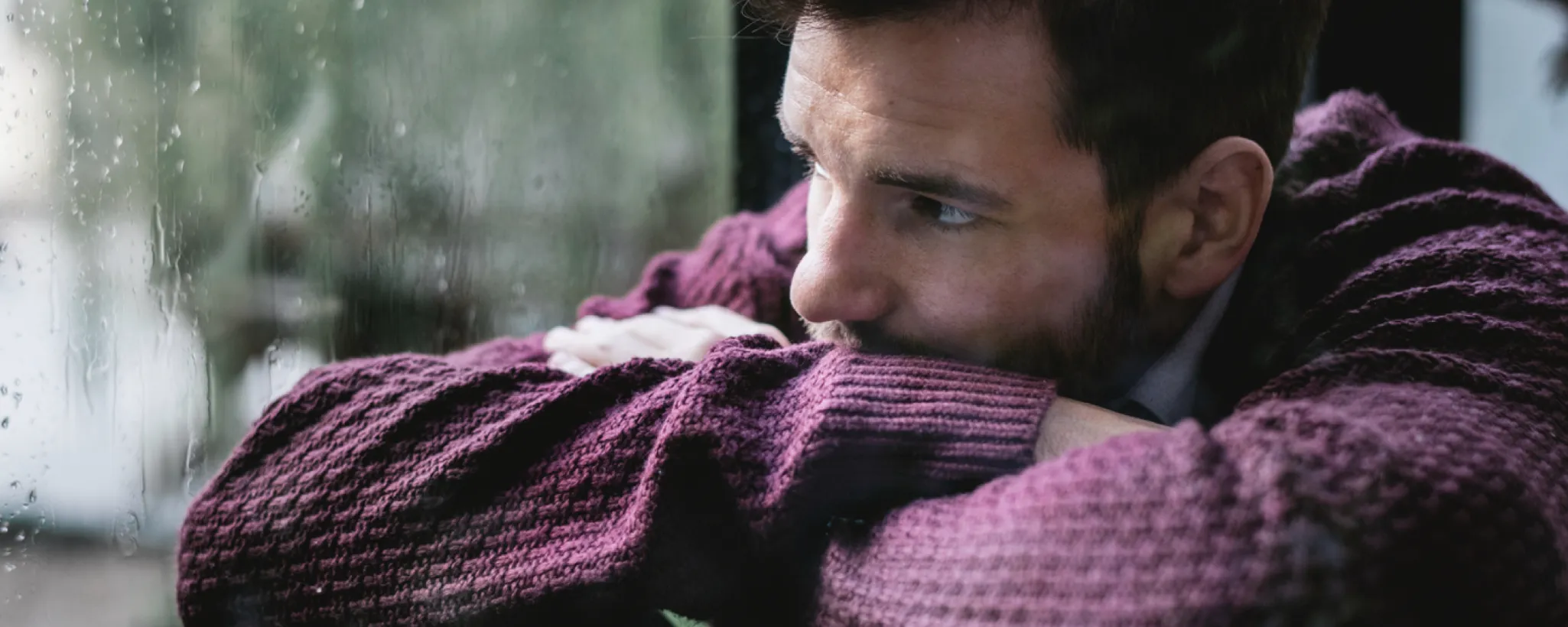 Ein Mann in einem weinroten Pullover lehnt mit verschränkten Armen an einem regennassen Fenster. Die trübe Atmosphäre vermittelt schlechte Laune und Depression.