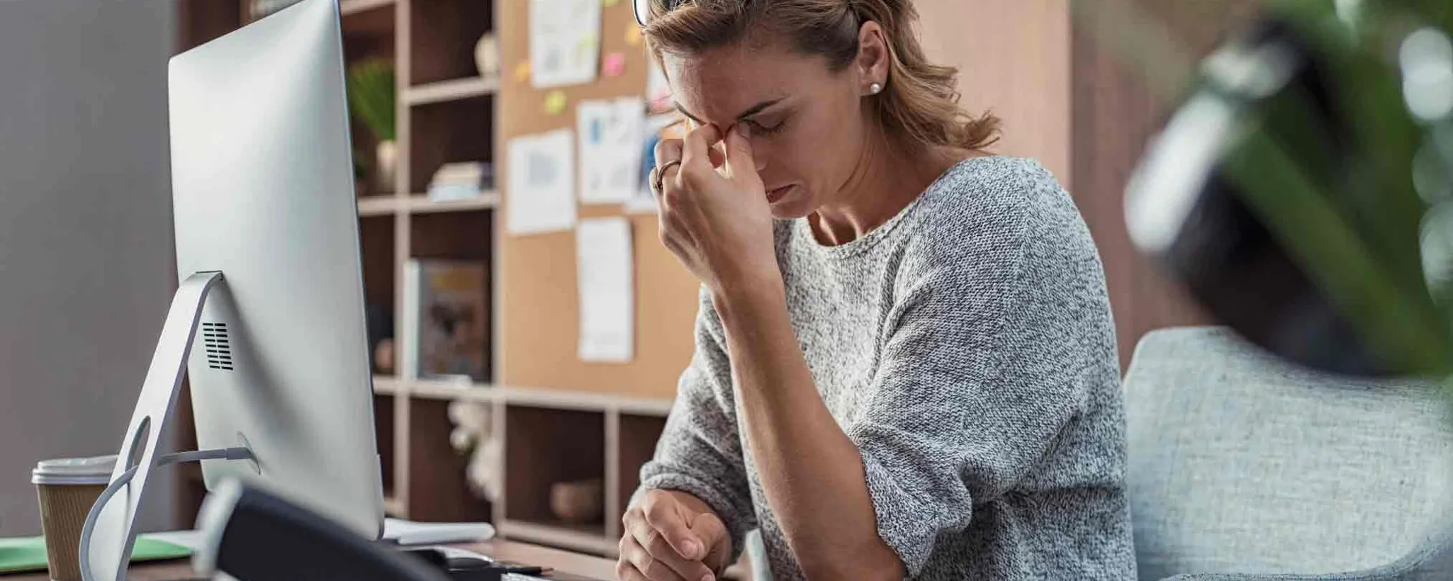 Eine Frau sitzt vor einem Computer in einer Büroumgebung und hält die Hand an die Nasenwurzel. Es entsteht der Eindruck einer belastenden Arbeitssituation.