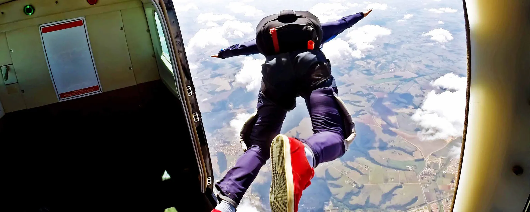 Ein Fallschirmspringer springt aus einem Flugzeug und schwebt in der Luft, während unter ihm eine Landschaft mit Feldern und Wolken sichtbar wird.