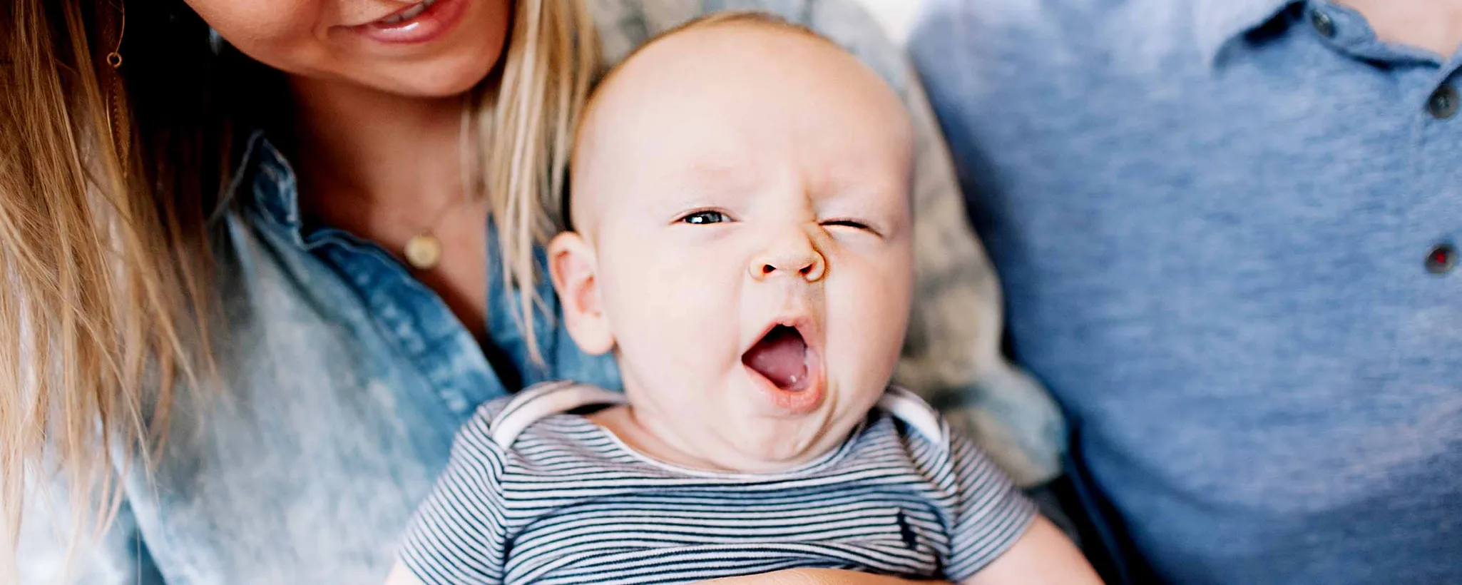 Ein Baby liegt auf dem Schoß seiner Eltern, die liebevoll und aufmerksam auf das Kind herabblicken. Beide tragen lässige Kleidung und sitzen dicht nebeneinander.