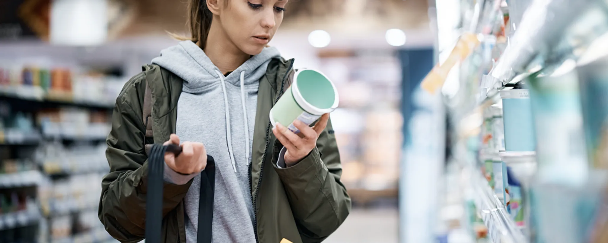Eine Person im Supermarkt hält eine Dose in der Hand und betrachtet sie aufmerksam, während sie mit der anderen Hand eine Einkaufstasche trägt, vermutlich um beim Einkaufen zu sparen. Die Regale sind mit verschiedenen Produkten gefüllt, die möglicherweise durch eine Versicherung geschützt sind.