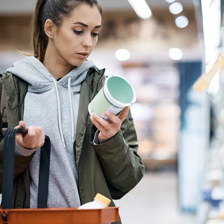 Eine Person im Supermarkt hält eine Dose in der Hand und betrachtet sie aufmerksam, während sie mit der anderen Hand eine Einkaufstasche trägt, vermutlich um beim Einkaufen zu sparen. Die Regale sind mit verschiedenen Produkten gefüllt, die möglicherweise durch eine Versicherung geschützt sind.