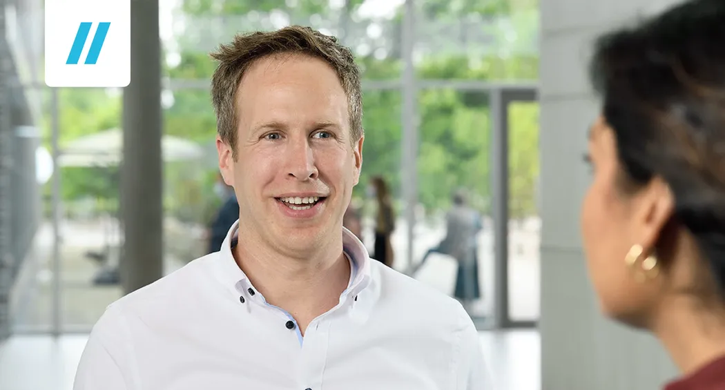 Robin Kazera steht in einem hellen, modernen Raum und führt ein Interview mit einer Person, die ihm gegenübersteht. Große Fenster lassen viel Tageslicht herein.
