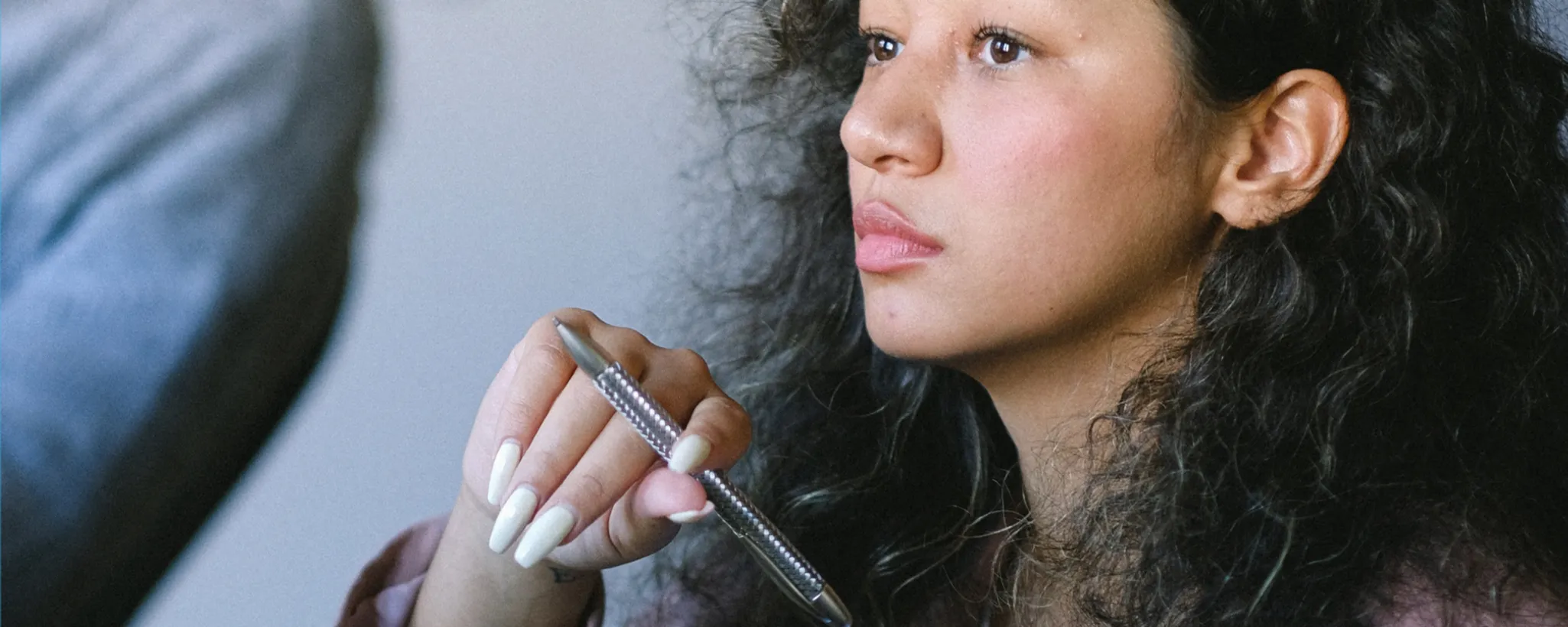 Eine Person mit lockigem Haar hält einen Stift in der Hand. Der Fokus liegt auf der Hand und dem Stift, was ein Bewusstsein für Details vermittelt. Im Hintergrund ist ein verschwommener Ärmel sichtbar, der an formelle Kleidung erinnert, wie sie oft in Versicherungen getragen wird.