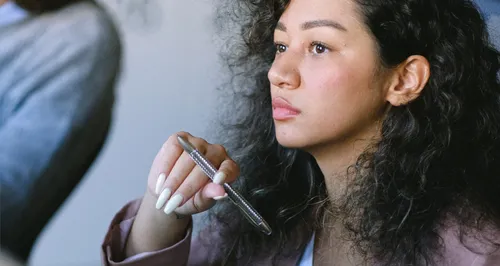 Eine Person mit lockigem Haar hält einen Stift in der Hand. Der Fokus liegt auf der Hand und dem Stift, was ein Bewusstsein für Details vermittelt. Im Hintergrund ist ein verschwommener Ärmel sichtbar, der an formelle Kleidung erinnert, wie sie oft in Versicherungen getragen wird.