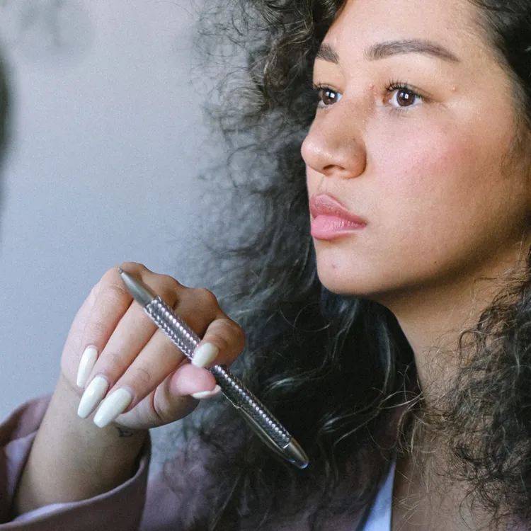 Eine Person mit lockigem Haar hält einen Stift in der Hand. Der Fokus liegt auf der Hand und dem Stift, was ein Bewusstsein für Details vermittelt. Im Hintergrund ist ein verschwommener Ärmel sichtbar, der an formelle Kleidung erinnert, wie sie oft in Versicherungen getragen wird.